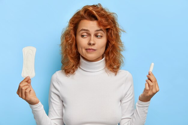 Women health care and hygiene concept. Indoor shot of hesitant young ginger woman holds two intimate products, chooses between tampon and pad during menses, thinks what gives better protection