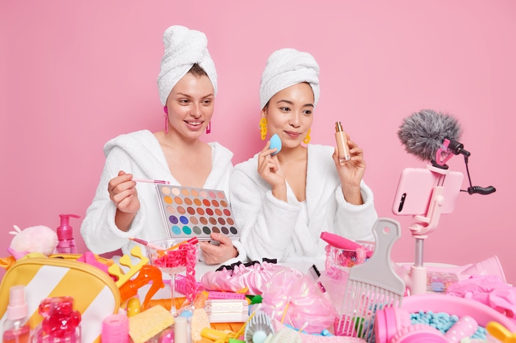 Free Photo women hold eyeshadow palette and bottle of foundation talk about doing professional makeup record online video have own blog dressed in bathrobes towels on head.