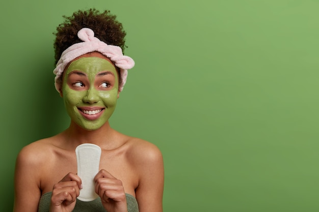 Women, hygiene, beauty, cosmetology concept. Happy pleased woman holds clean sanitary napkin for using during menstruation, looks with toothy smile on right side, isolated on green wall