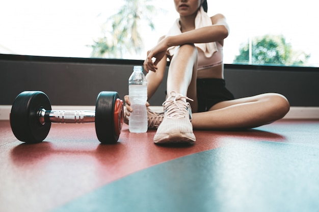 女性は運動後、座ってリラックスします。水筒とダンベルがあります。