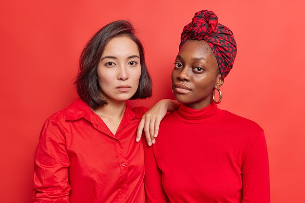 women stand closely to each other have calm confident look at camera dressed in red clothing have natural beauty healthy skin pose in studio. Diverse lesbian females