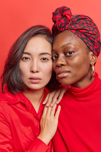 빨간 옷을 입고 카메라를 보고 자신감 넘치는 표정으로 서로 가까이 서 있는 여성들은 좋은 관계를 유지하고 있습니다. 혼혈 여성 모델이 실내에서 포즈를 취하고 있습니다. 다양성 개념