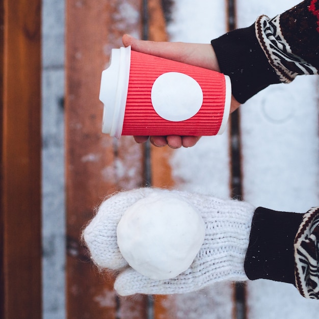 Womn tiene la palla di neve a mano in guanto bianco e tazza di carta rossa in un altro