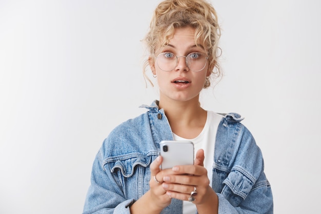 Wondered amazed cute blond woman messy curly bun wearing glasses denim jacket playing cool smartphone game holding phone searching address using phone app turn camera questioned surprised