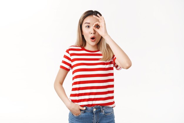 Wondered amused emotive blond asian woman drop jaw folding lips wow sound stare camera excited amazed show okay ok monocle gesture astonished look through fingers white background