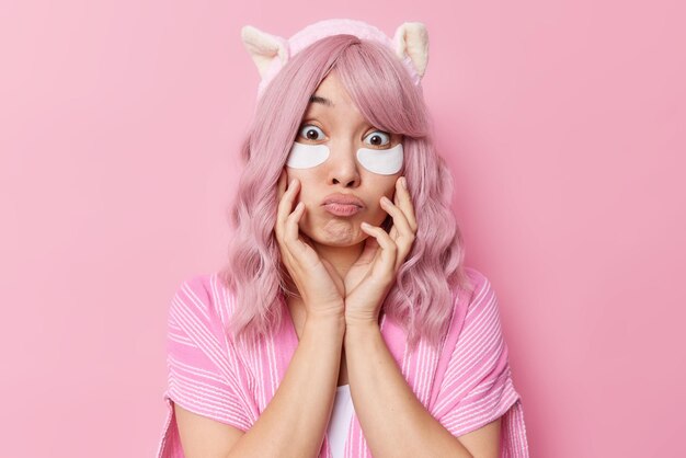 Wondered Asian woman looks surprisingy at camera has bugged eyes lips rounded applies beauty patches under eyes to remove wrinkles dressed casually isolated over pink background Skin care concept