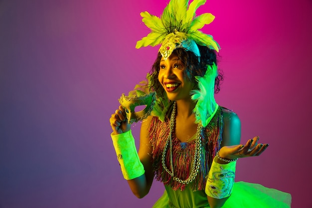 Wondered. Beautiful young woman in carnival, stylish masquerade costume with feathers dancing on gradient wall in neon. Concept of holidays celebration, festive time, dance, party, having fun.