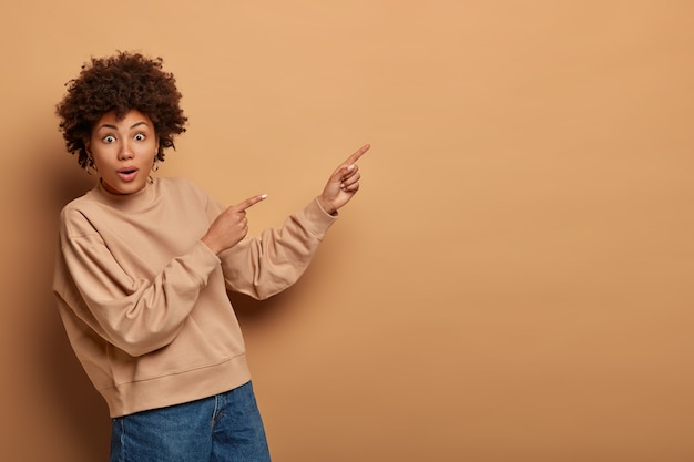 wondered curly haired woman has curious excited gaze, shows product on empty space, gives advice, dressed in brown sweatshirt, says click on link, isolated on beige wall