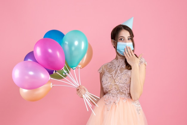 wondered girl with party cap putting hand on mouth holding colorful balloons on pink