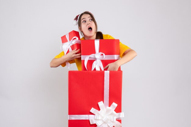 白の大きなクリスマスプレゼントの後ろに立っている贈り物を保持しているサンタの帽子を持つ不思議な女の子