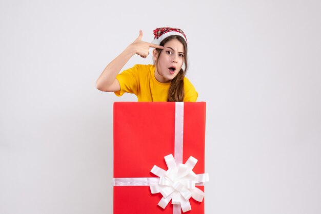 wondered girl with santa hat keeping finger gun to her temple standing behind big xmas gift on white