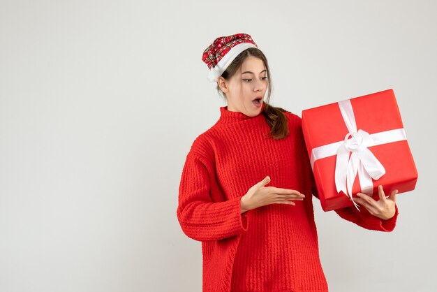 удивленная девушка в новогодней шапке, указывая на свой рождественский подарок на белом