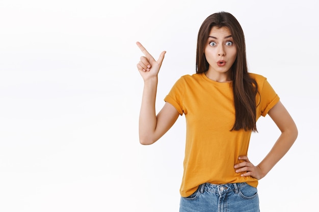 Wondered, impressed good-looking woman in yellow t-shirt, folding lips amazed, say wow and stare camera astonished as discuss new product, pointing upper left corner, checking out cool promo