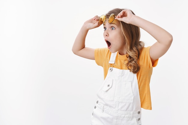 Wondered impressed little gorgeous girl, child travel abroad parents look fascinated turn left ambushed, take-off glasses speechless and astonished, seeing awesome offer, white wall