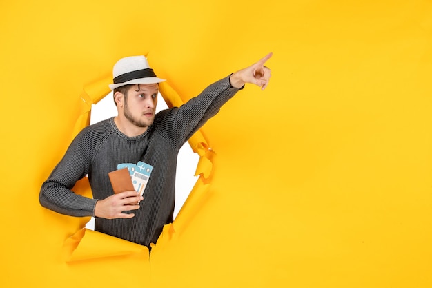 Бесплатное фото Удивленный человек в шляпе держит заграничный паспорт с билетом и указывает что-то в разорванной на желтой стене