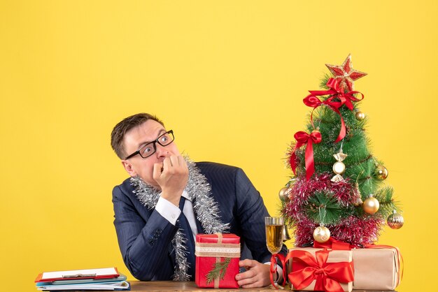 wondered man with eyeglasses sitting at the table near xmas tree and presents on yellow