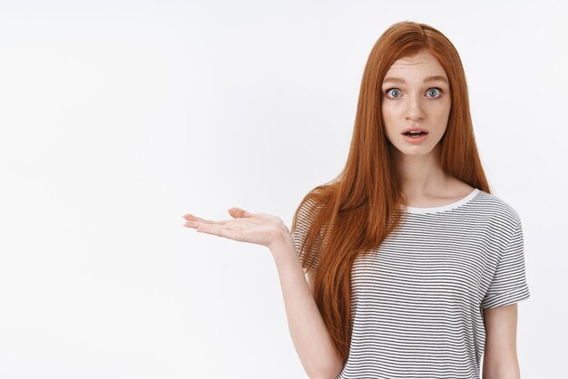 Wondered questioned attractive young european redhead girl blue eyes open mouth gasping impressed raise palm holding object product device hand reacting speechless amazed white background