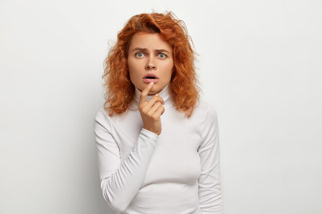 Wondered surprised young woman looks attentively with opened mouth , reacts on something astonishing, has ginger hair, green eyes, wears casual turtleneck, isolated on white wall