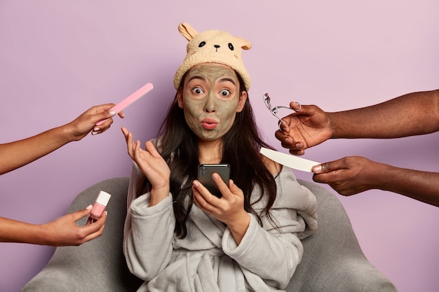 Wondered woman with shocked expression, uses mobile phone during beauty treatment