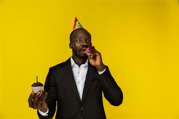 Wondered young afroamerican guy in black suit and birthday hat with burning candle on the cupcake 