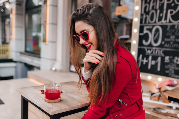 Замечательная брюнетка в красном позирует с застенчивой улыбкой в уличном кафе