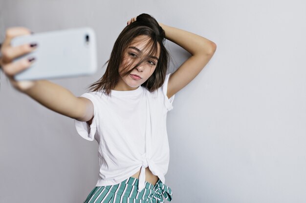 Wonderful brunette girl touching her hair while taking picture of herself. Indoor shot of pretty woman using phone for selfie.