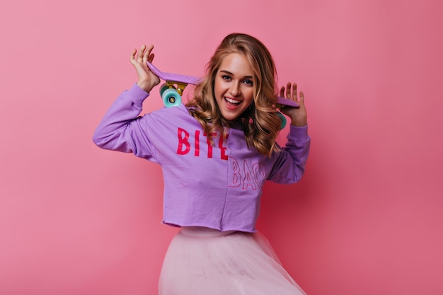 Wonderful caucasian girl posing with cheerful smile on pastel. Fashionable blonde lady with skateboard expressing happiness.