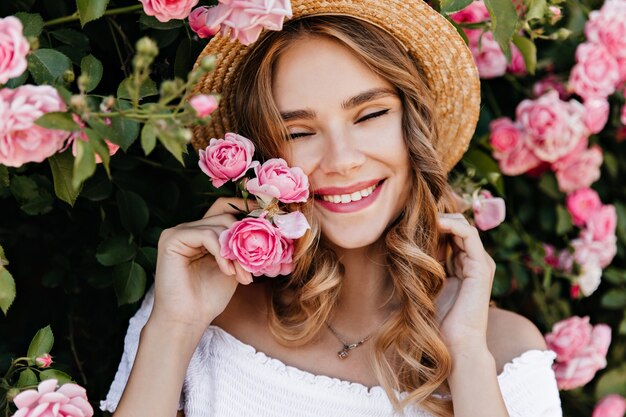 행복을 표현하는 밀 짚 모자에 멋진 백인 여자. 여름 날에 장미 덤불 근처에 서있는 귀여운 여성 모델.