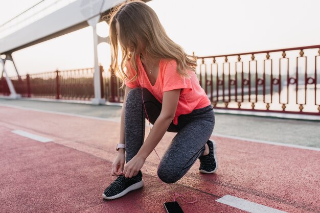 Wonderful caucasian lady in earphones ties up her shoelaces at stadium. Outdoor shot of cute girl sitting at cinder track and listening music.