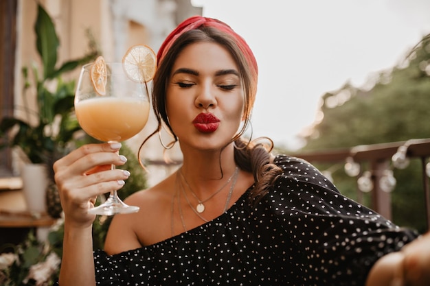 Замечательная крутая женщина с красными губами, круглыми серьгами и стильной повязкой на голове в современном наряде в горошек, дует воздушный поцелуй и держит стакан апельсинового коктейля
