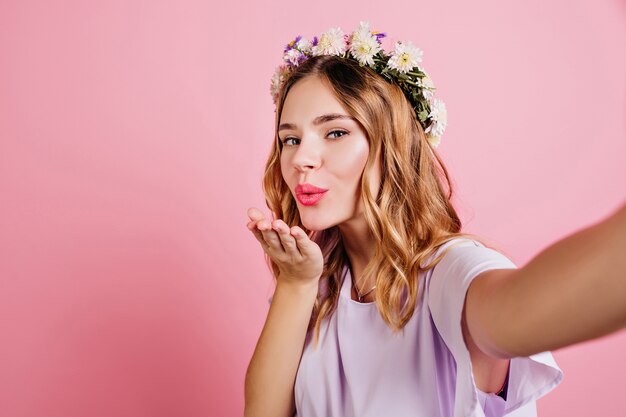 Wonderful curly woman in circlet of flowers sending air kiss to camera