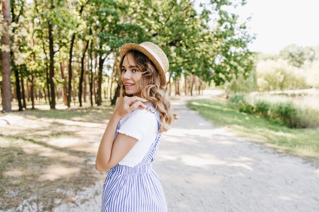 サマーパークを歩いて笑っている素敵な金髪の女の子。自然に喜んでポーズをとる麦わら帽子の愛らしい女性の屋外写真。