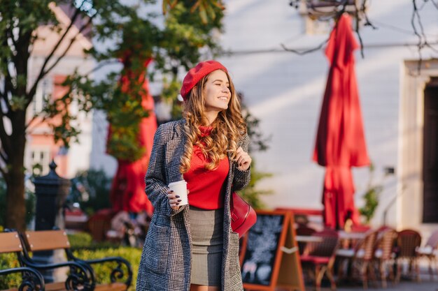 Wonderful female model in gray clothes walking down the street with cup of coffee