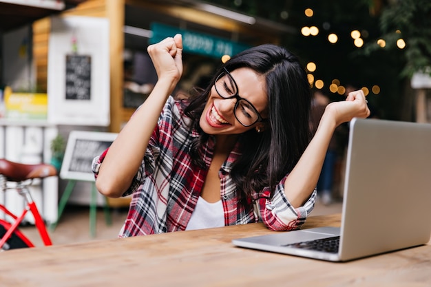 Wonderful female student sitting in cafe with laptop and laughing. Pleasant freelancer in glasses expressing happiness.