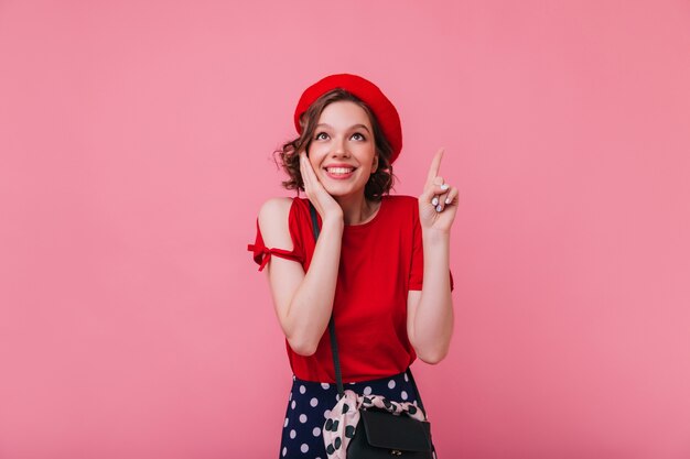 Wonderful french girl with wavy hairstyle posing with surprised smile. Indoor photo of graceful white woman in red beret isolated.