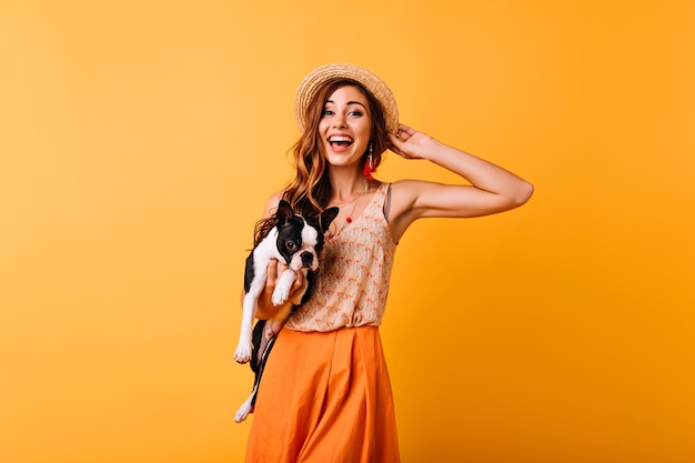 Wonderful ginger girl in summer hat expressing happiness during portraitshoot with dog. Amazing pretty girl holding bulldog and smiling.