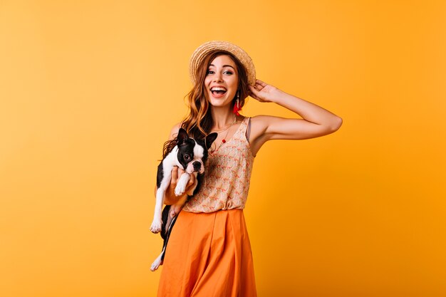 Замечательная рыжая девушка в летней шляпе, выражая счастье во время портретной съемки с собакой. Удивительная красивая девушка держит бульдога и улыбается.