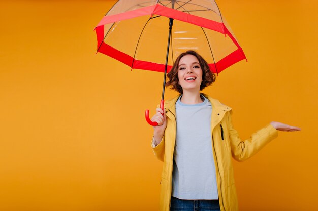 Wonderful girl in good mood laughing while posing with red umbrella. Indoor photo of trendy caucasian lady with sparkle makeup enjoying photoshoot with parasol.