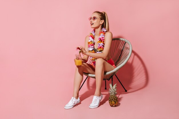Wonderful girl with blonde hair in swimsuit, sunglasses and necklace of flowers sitting on chair and holding cocktail on pink wall