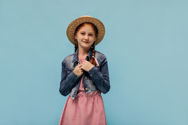 Wonderful girl with freckles and red hair in denim jacket, cool hat and trendy dress looking at front on blue wall