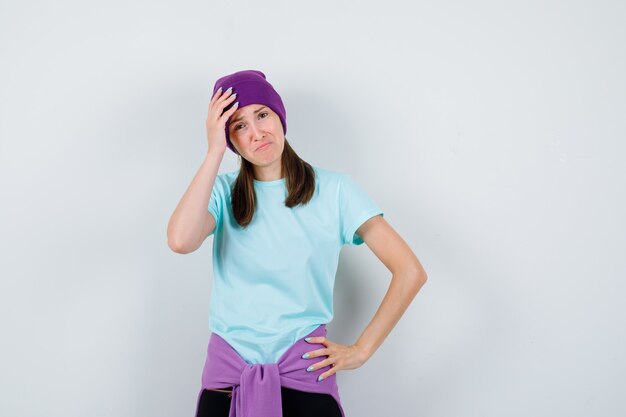 Wonderful lady in blouse, beanie keeping hand on head and looking mourned , front view.