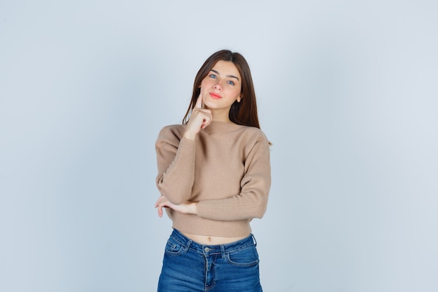 Wonderful lady keeping finger on cheek in sweater, jeans and looking charming , front view.