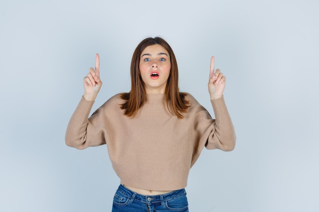 Free photo wonderful lady pointing up and opening mouth in sweater, jeans and looking shocked. front view.