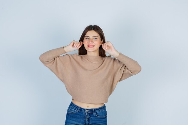 Wonderful lady pulling her earlobes while smiling in sweater, jeans and looking joyful. front view.