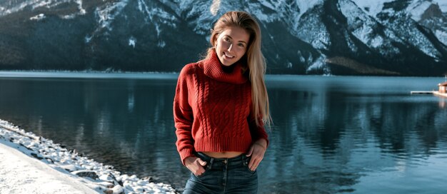 深い湖と素晴らしい山の景色の雪に覆われた海岸に屋外に立っている素晴らしい女性。特大のセーターとジーンズの陽気な女の子。化粧なしと長いブロンドの髪型。青い澄んだ空。