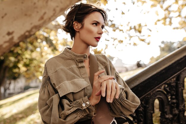 Wonderful lady in wide denim jacket looking away outside. Trendy woman with short curly hairstyle and bright lips posing outdoors.