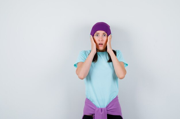 Wonderful lady with hands on head in blouse, beanie and looking terrified , front view.