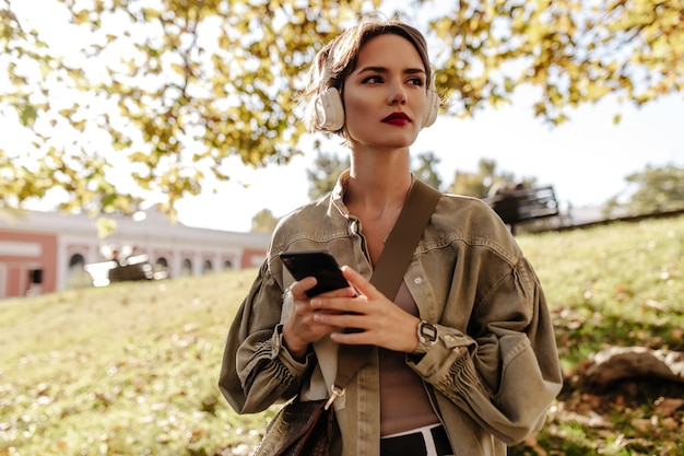 Wonderful lady with short hair in white headphones and olive jacket looking away outdoors. woman with handbag holds phone outside.