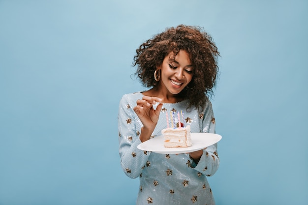 Free photo wonderful lady with wavy stylish hairstyle in earrings and blue shiny dress smiling and holding piece of cake with candles on blue wall..