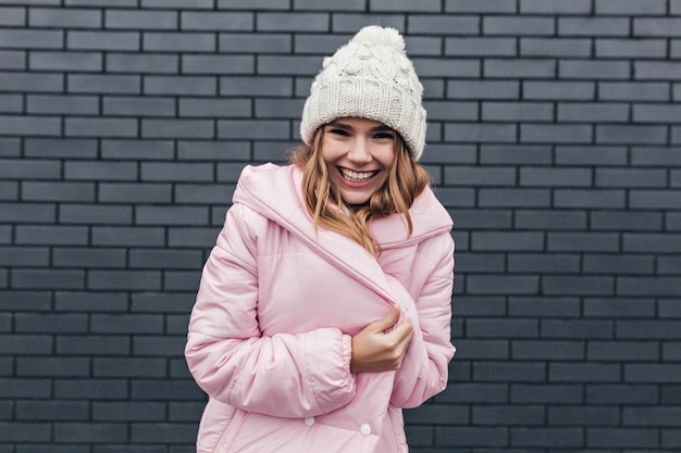 Wonderful laughing girl posing in pink coat. Outdoor photo of excited blonde lady in trendy winter hat.